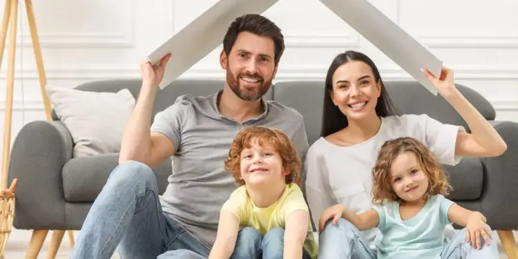 Familia sonriente sosteniendo un tejado de cartón simulando una casa en la sala de estar