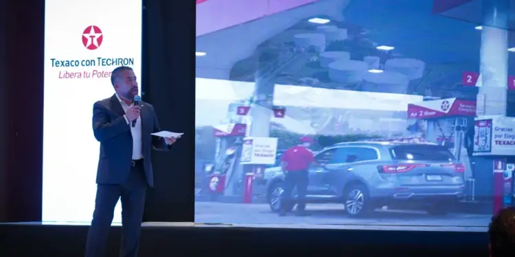 Un ponente en traje formal hablando en un escenario con una gran pantalla que muestra una estación de servicio y vehículos.