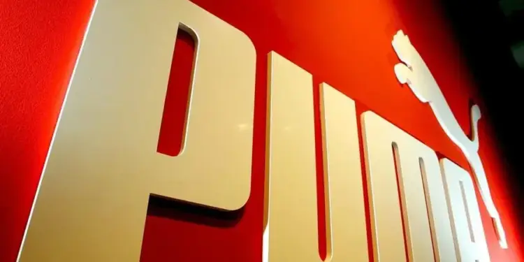 Primer plano del logo tridimensional de PUMA en color dorado sobre un fondo rojo brillante, destacando el felino saltando y las letras de la marca.