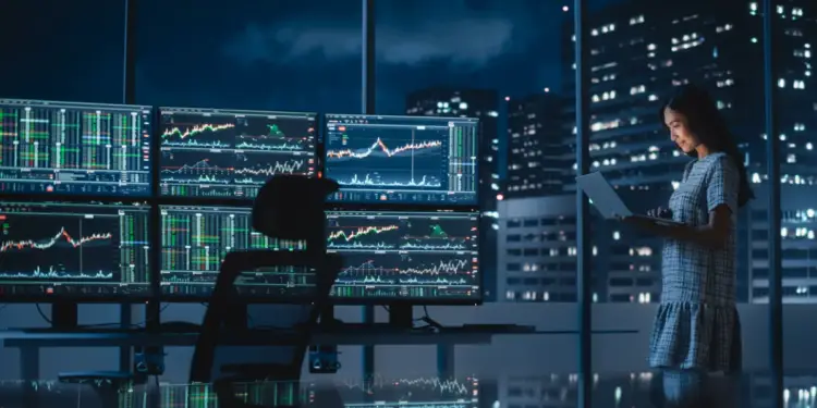 Una mujer de pie en una oficina nocturna frente a múltiples pantallas con gráficos financieros y una ciudad iluminada de fondo.