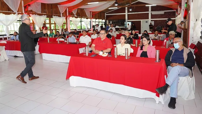 Un expositor habla frente a una audiencia sentada en mesas con manteles rojos durante una conferencia empresarial.