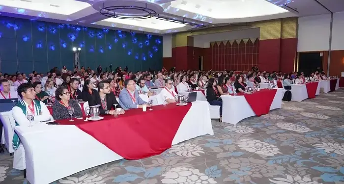 Numerosos asistentes sentados en mesas con manteles blancos y rojos durante una conferencia empresarial en un gran salón.