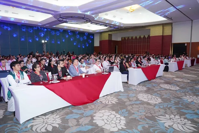 Numerosos asistentes sentados en mesas con manteles blancos y rojos durante una conferencia empresarial en un gran salón.