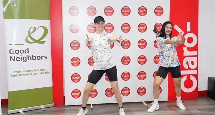 Dos mujeres bailando y haciendo ejercicio frente a un panel con logotipos de marcas corporativas en un evento.