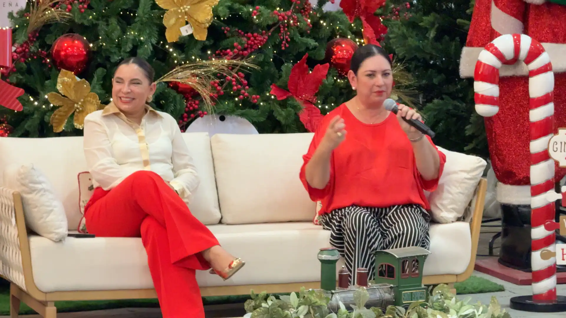 Dos mujeres sentadas en un sofá blanco conversando en un evento corporativo navideño con decoración festiva de fondo.