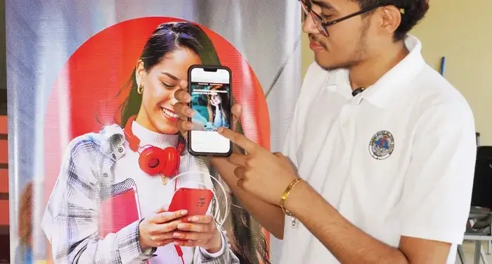 Joven con camisa blanca y gafas señalando la pantalla de un teléfono móvil frente a un cartel publicitario.