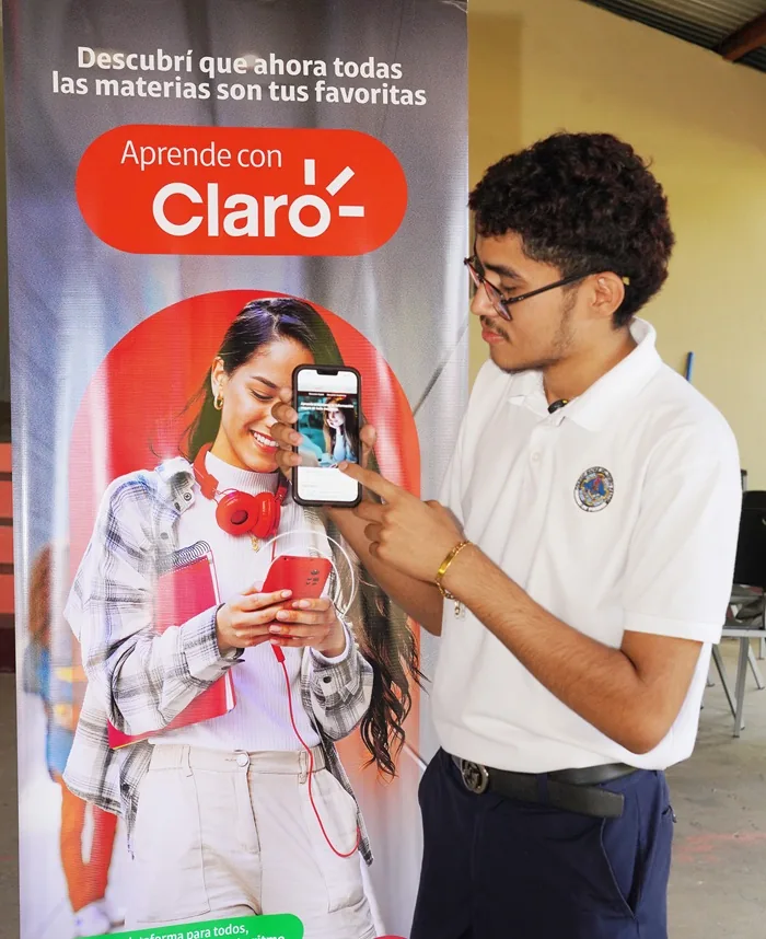 Joven con camisa blanca y gafas señalando la pantalla de un teléfono móvil frente a un cartel publicitario.