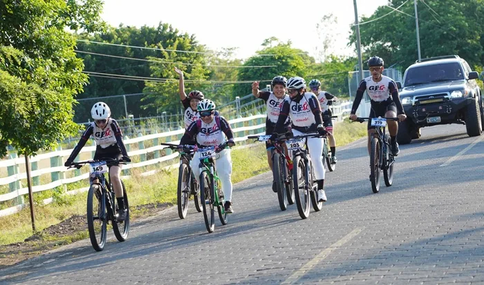 Grupo de personas con equipo deportivo y cascos montando bicicletas en una carretera pavimentada rodeada de vegetación.