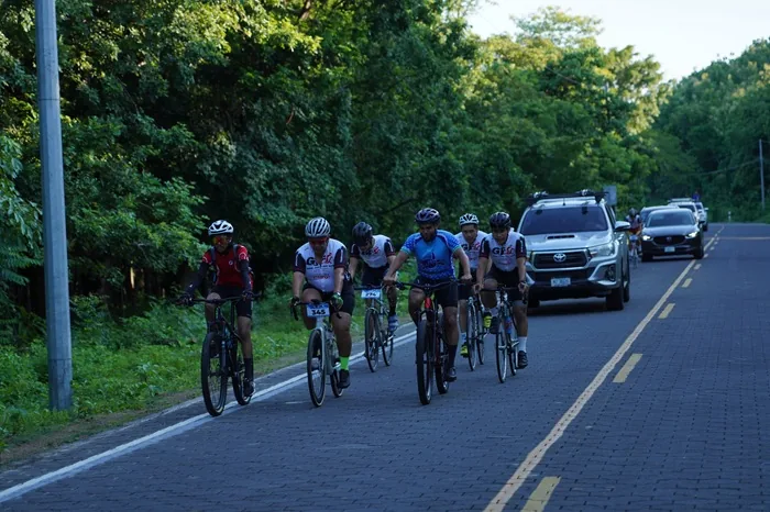 Un grupo de ciclistas pedaleando en fila por una carretera asfaltada rodeada de vegetación, con vehículos detrás.