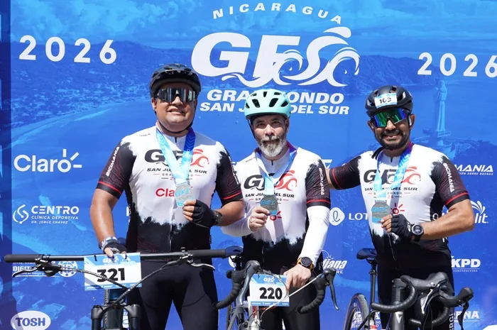 Tres ciclistas posan sonrientes con medallas y bicicletas frente a un cartel azul del evento Gran Fondo San Juan del Sur.
