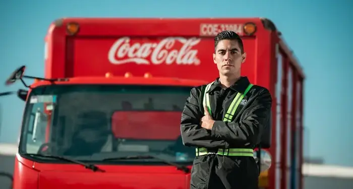 Hombre con uniforme oscuro y chaleco reflectante de seguridad, brazos cruzados, de pie frente a un camión de reparto rojo con logos Coca-Cola e Isuzu.