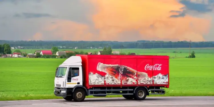 Camión de reparto con la marca Coca-Cola, transportando productos a través de un paisaje rural con campos verdes y un cielo nublado.