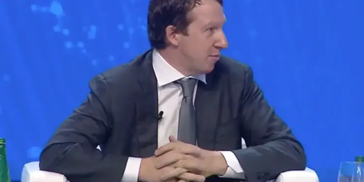 Hombre ejecutivo en traje gris y corbata, sentado en silla blanca con manos entrelazadas, en un evento profesional o conferencia de negocios con fondo azul.