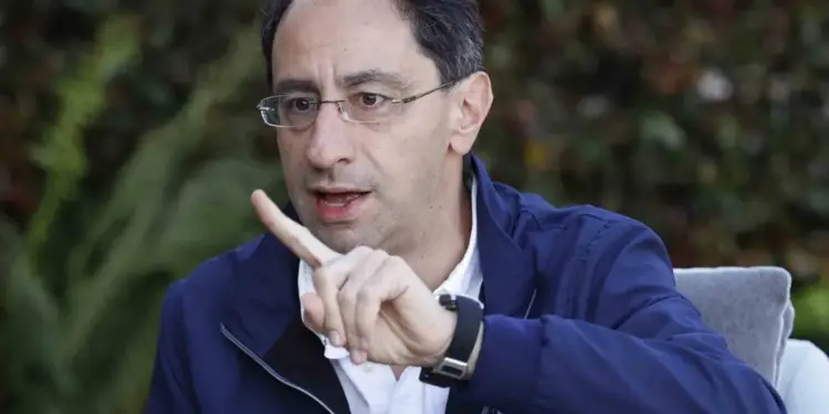 Hombre con gafas y chaqueta azul oscuro, gesticulando con el dedo índice levantado. En su chaqueta se lee 'FIRME POR LA PATRIA' y 'PRESIDENTE'. Lleva un micrófono y un reloj.