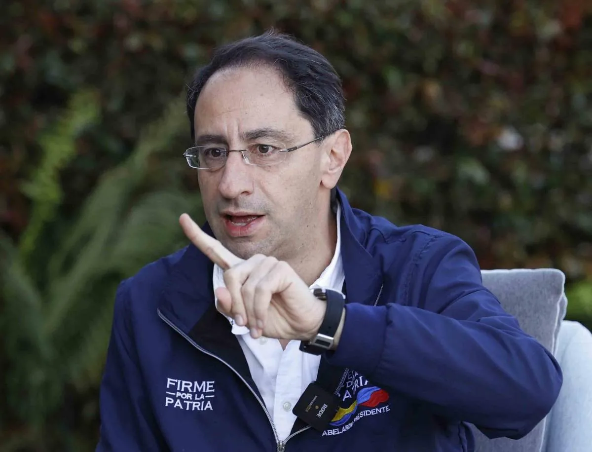 Hombre con gafas y chaqueta azul oscuro, gesticulando con el dedo índice levantado. En su chaqueta se lee 'FIRME POR LA PATRIA' y 'PRESIDENTE'. Lleva un micrófono y un reloj.
