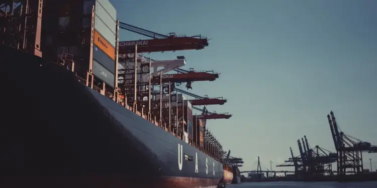 Gran buque portacontenedores con la inscripción 'UASC' en su casco, navegando en un puerto. Múltiples contenedores y grúas portuarias con 'BURCHARDKAI' son visibles al fondo.
