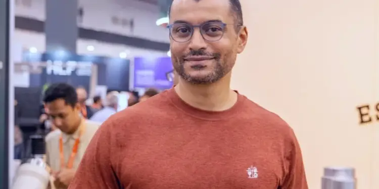 Hombre sonriendo a la cámara, viste una camiseta de color terracota y gafas. Al fondo se observan personas y stands de un evento empresarial.