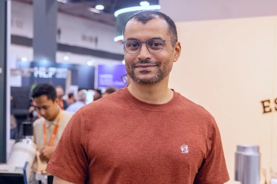 Hombre sonriendo a la cámara, viste una camiseta de color terracota y gafas. Al fondo se observan personas y stands de un evento empresarial.
