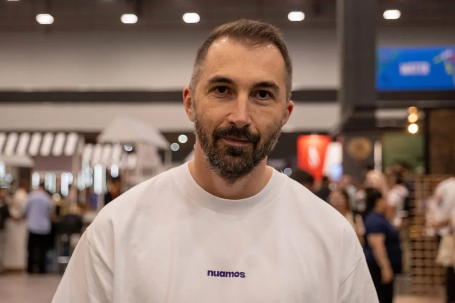 Primer plano de un hombre con barba corta y camiseta blanca con el logo "nuamos" en letras moradas, en un evento con gente y stands al fondo.