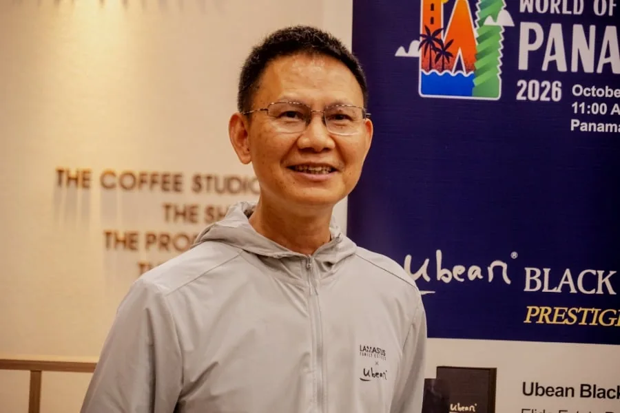 Profesional asiático sonriente con gafas y chaqueta gris, frente a un banner azul de "World of Panama 2026" y "Ubean Black Prestige".