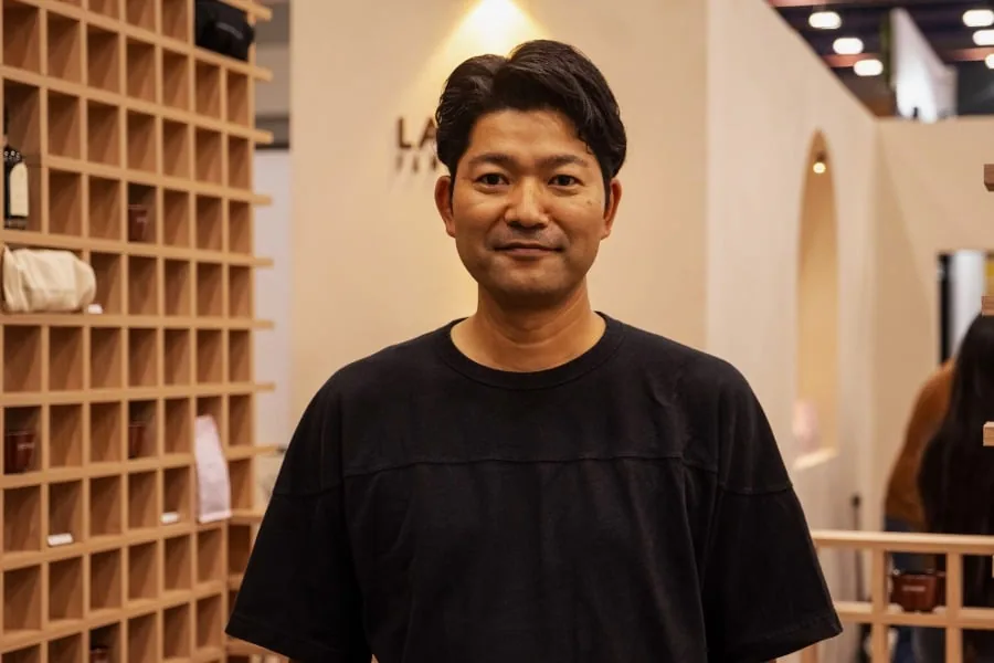 Retrato de un hombre asiático sonriendo en un entorno comercial con estanterías de madera y productos exhibidos.