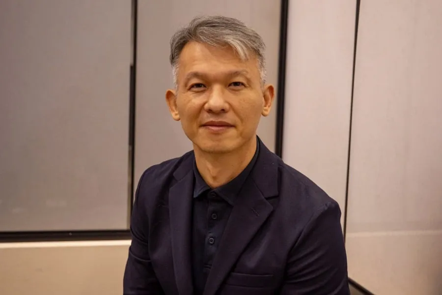 Retrato de un hombre de mediana edad, de rasgos asiáticos, con cabello gris y oscuro, vistiendo un blazer y polo azul marino. Mira directamente a la cámara con expresión seria en un entorno profesional.