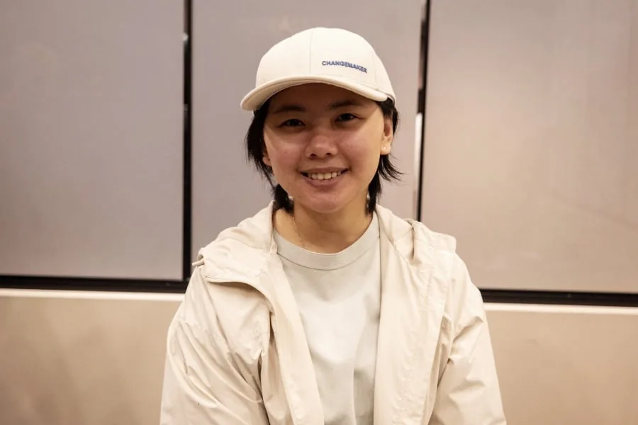 Persona joven sonriendo con chaqueta clara y gorra blanca que dice CHANGEMAKER sobre fondo neutro.