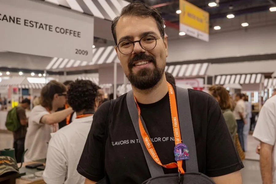 Hombre sonriente con gafas y acreditación naranja en una feria comercial de café rodeado de asistentes.