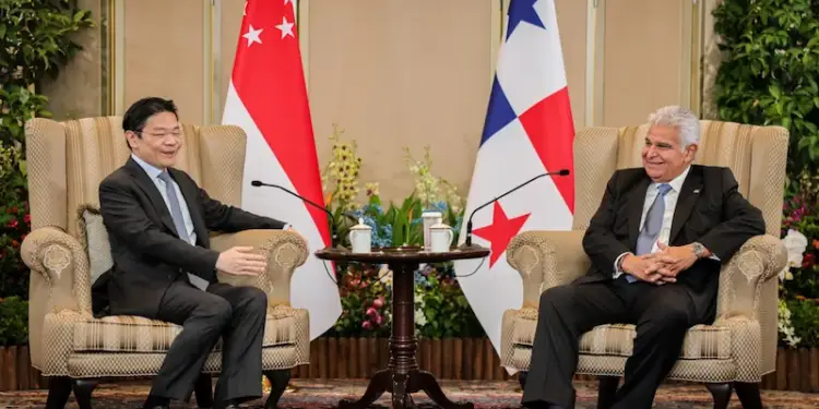 Dos hombres en traje sentados en sillones conversando frente a dos banderas de diferentes países.