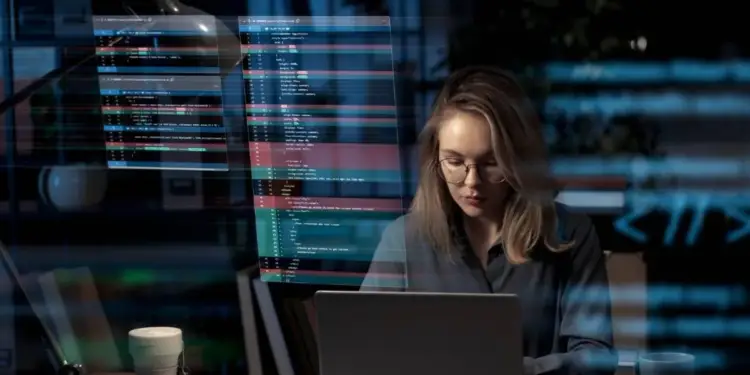 Mujer profesional con gafas trabajando en un portátil, con pantallas de código superpuestas. Representa desarrollo tecnológico y análisis de datos.