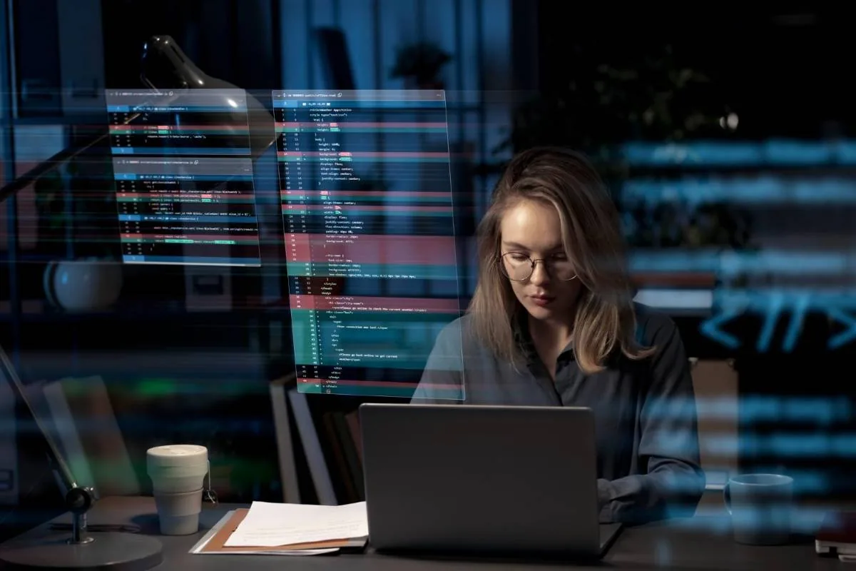 Mujer profesional con gafas trabajando en un portátil, con pantallas de código superpuestas. Representa desarrollo tecnológico y análisis de datos.