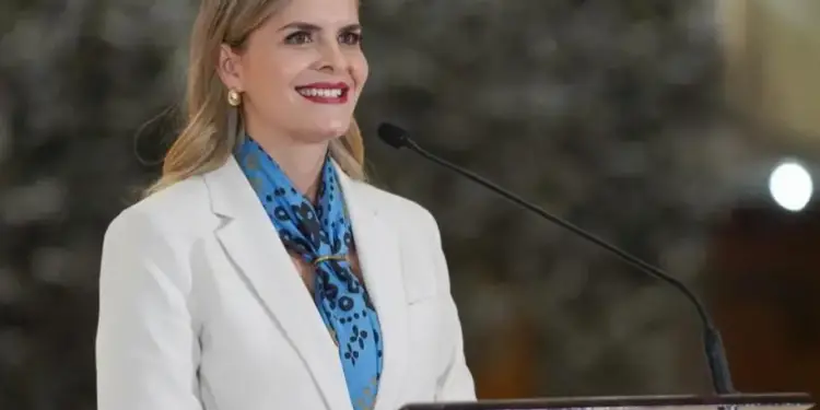 Oradora en evento de negocios, con blazer blanco y pañuelo azul, sonríe mientras habla en un podio con micrófono.