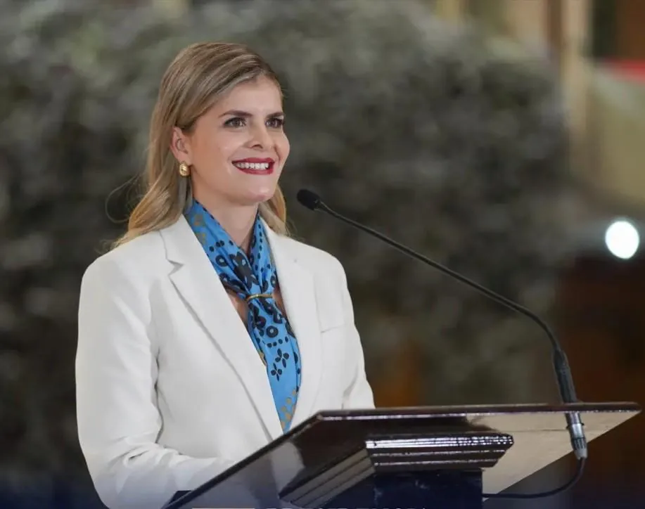 Oradora en evento de negocios, con blazer blanco y pañuelo azul, sonríe mientras habla en un podio con micrófono.