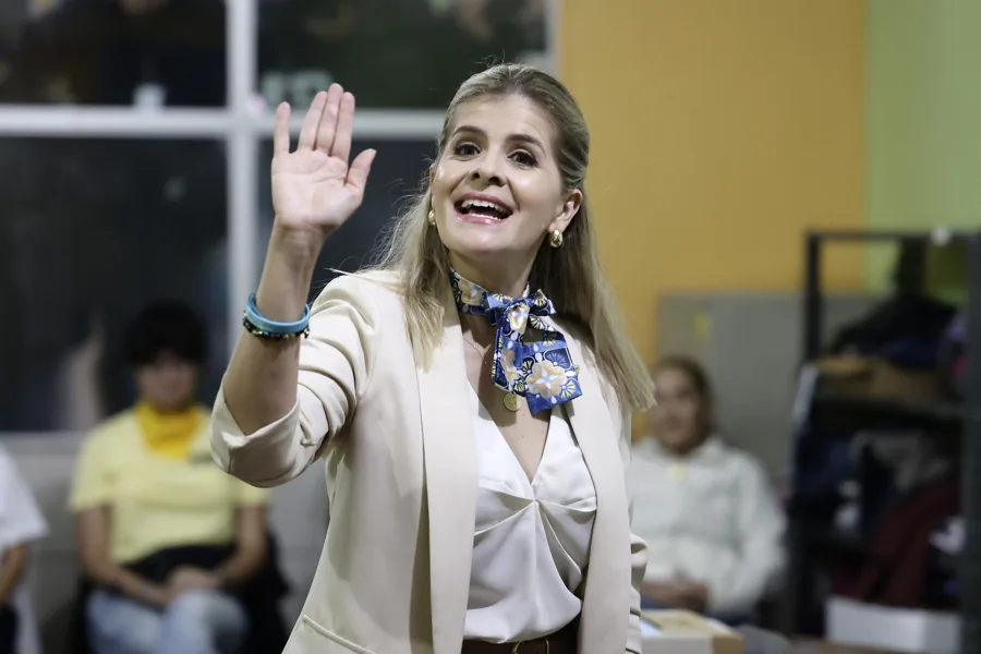 Mujer rubia con chaqueta beige y pañuelo saludando con la mano sonriente en un entorno de oficina o sala de reuniones.
