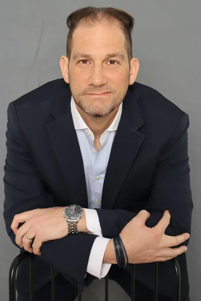 Hombre de negocios sonriente con chaqueta oscura, camisa a rayas y reloj de pulsera frente a fondo gris