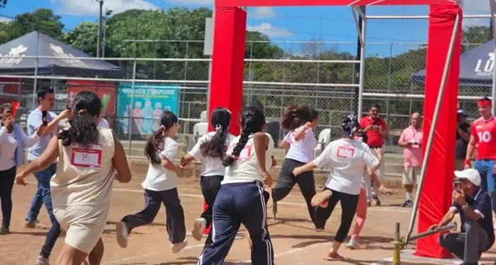 Niños y jóvenes corren hacia un arco rojo de meta con un cerdito de ahorro, simbolizando el logro de objetivos financieros en un evento.