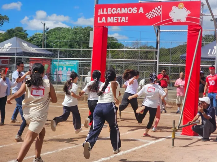 Niños y jóvenes corren hacia un arco rojo de meta con un cerdito de ahorro, simbolizando el logro de objetivos financieros en un evento.