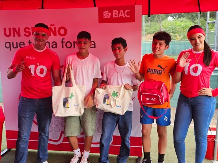 Cinco personas, incluyendo adultos y jóvenes, posan en un evento patrocinado por BAC y Kash. Muestran bolsas y una mochila con logos.