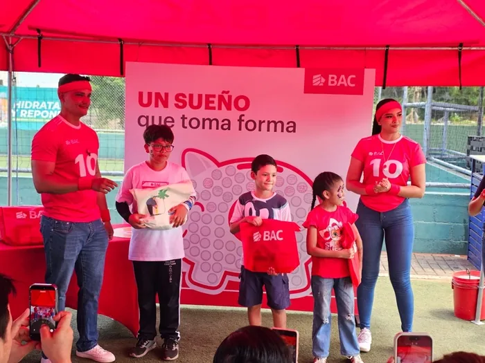 Adultos y niños en un evento de BAC bajo un toldo rojo, con un banner de "UN SUEÑO que toma forma" y una alcancía. Los niños reciben obsequios.