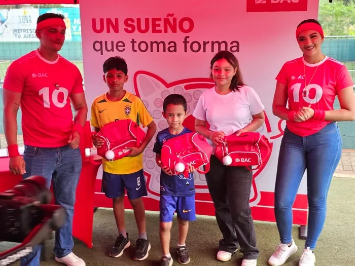 Cinco personas, dos adultos y tres niños, posan frente a un banner rojo de BAC. Los niños sostienen bolsas rojas con el logo de BAC.