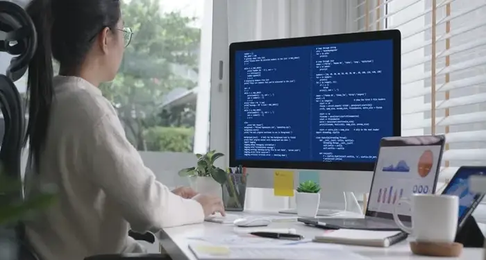 Mujer sentada frente a un monitor con código y una laptop con gráficos financieros en un escritorio