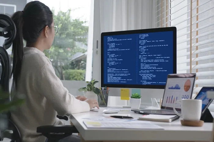 Mujer sentada frente a un monitor con código y una laptop con gráficos financieros en un escritorio