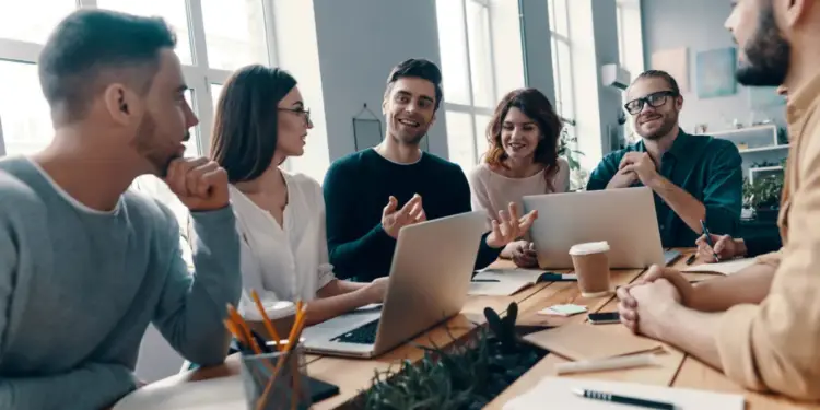 Un grupo diverso de profesionales jóvenes colaborando activamente alrededor de una mesa de madera, discutiendo ideas con laptops y cuadernos abiertos.