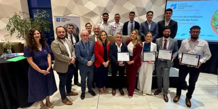 Grupo de personas posando para una foto grupal, algunos sosteniendo certificados de premiación, con un fondo de pantalla que muestra el logo de Luxaid Challenge Fund.