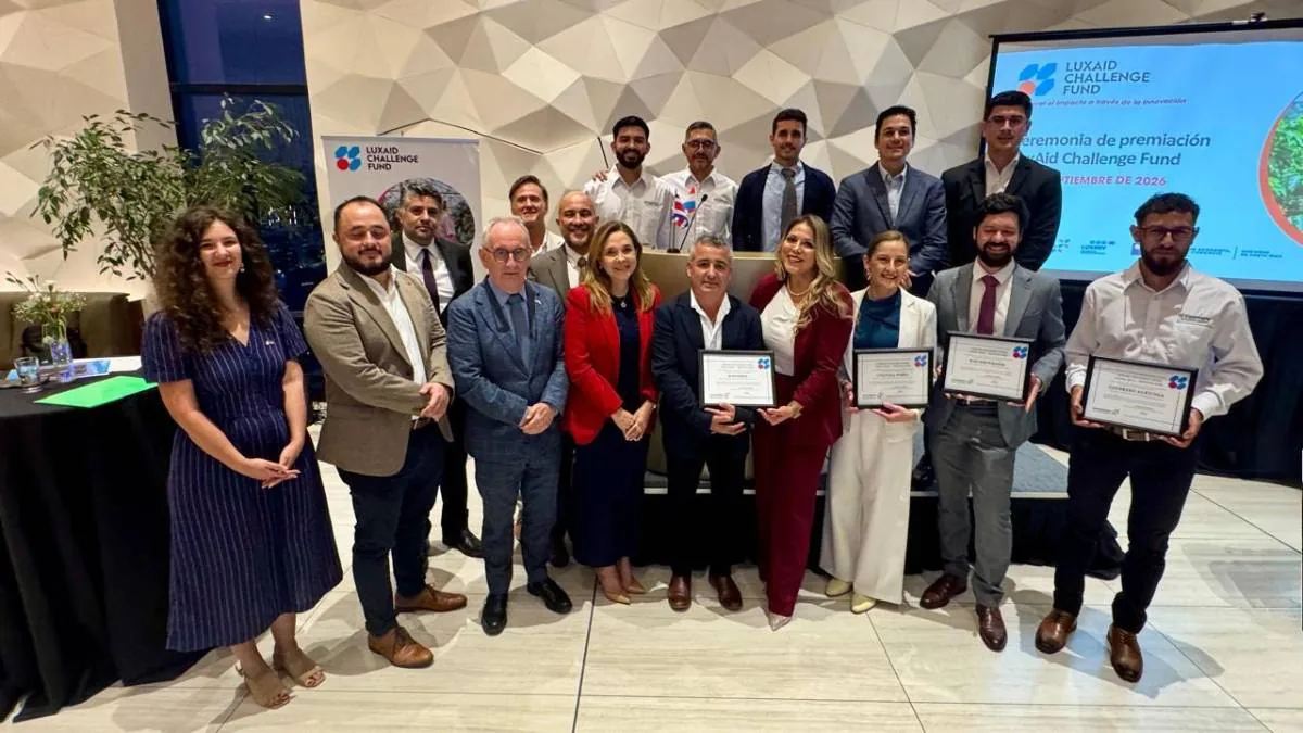 Grupo de personas posando para una foto grupal, algunos sosteniendo certificados de premiación, con un fondo de pantalla que muestra el logo de Luxaid Challenge Fund.