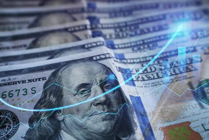 Primer plano de billetes de cien dólares estadounidenses con un gráfico financiero azul brillante superpuesto, mostrando una tendencia alcista y barras de crecimiento.