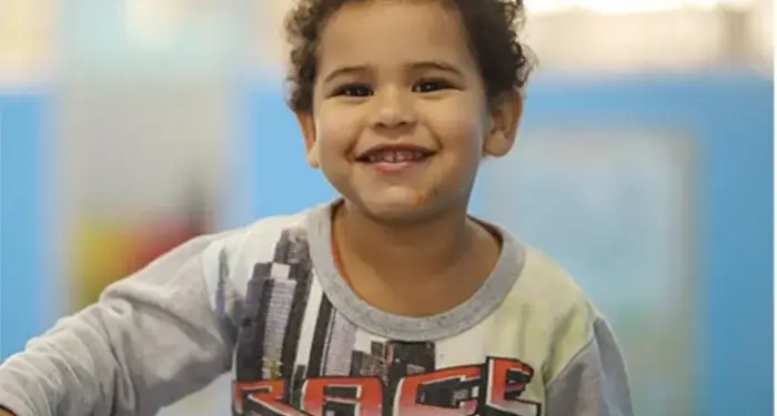Un niño pequeño con cabello rizado sonriendo alegremente a la cámara en una habitación con paredes azules.