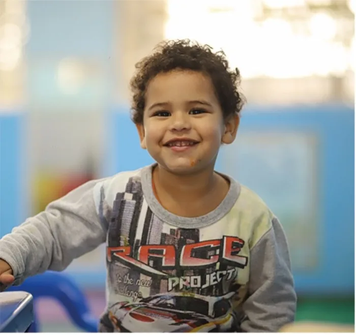 Un niño pequeño con cabello rizado sonriendo alegremente a la cámara en una habitación con paredes azules.