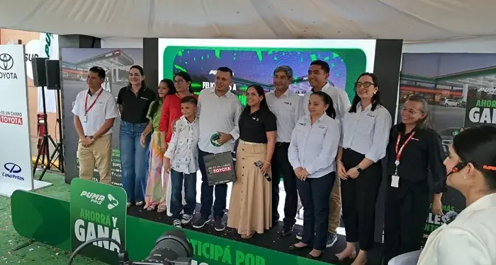 Grupo de hombres y mujeres de pie sonriendo en un escenario frente a una pantalla con un letrero verde.
