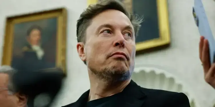Retrato de Elon Musk vistiendo traje oscuro con cuadros al fondo y una mano sosteniendo un teléfono móvil.